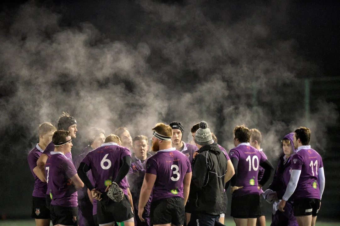 A group of male rugby players wearing purple shirts having a team talk on the pitch at night time. They look like they are steaming.