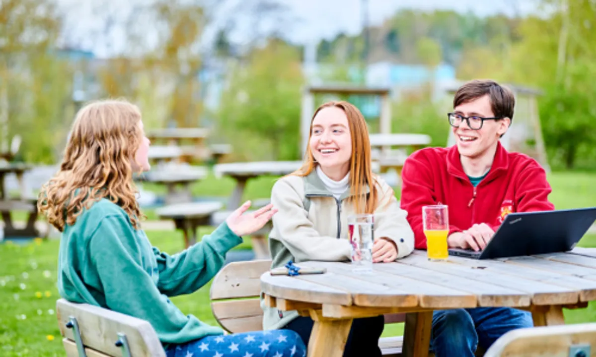 Three students sat outside on a bench with drinks and a laptop