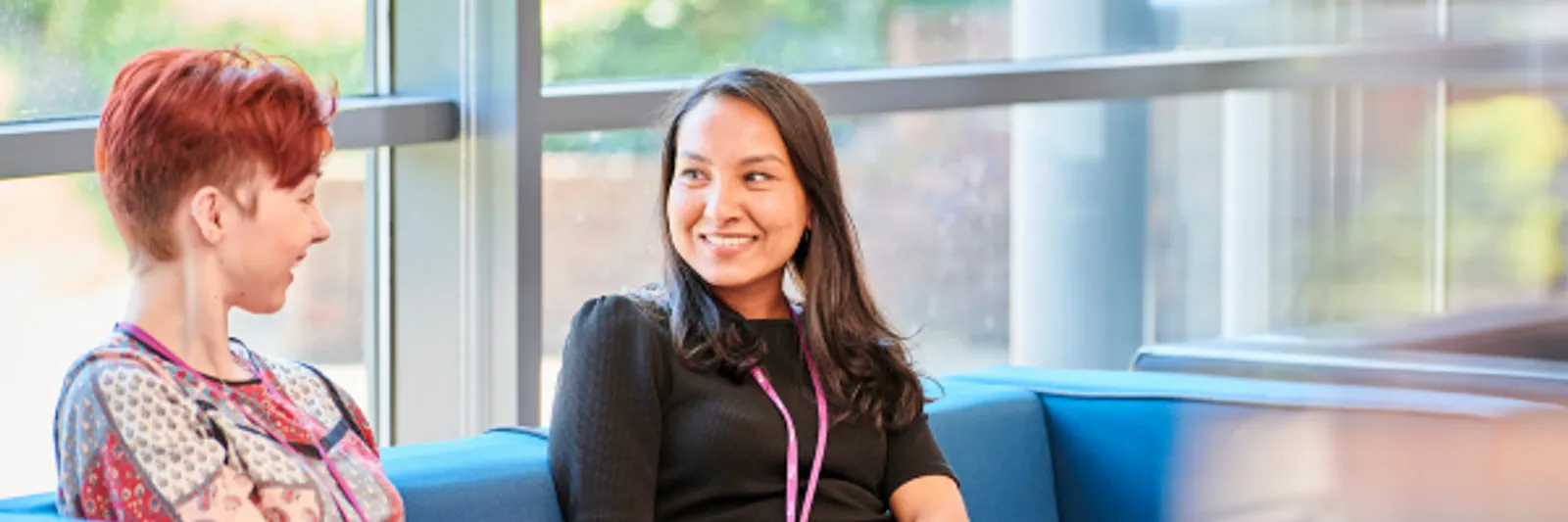 Two women sit on a low sofa in a brightly lit, modern space. A window behind them shows that it's sunny outside with pale green trees visible. The woman on the right has long black hair and wears a black dress. She has a Durham University lanyard on a purple ribbon. The woman on the left has short, dark auburn hair and her nose and ears are pierced. She wears a brightly patterned top. They work t