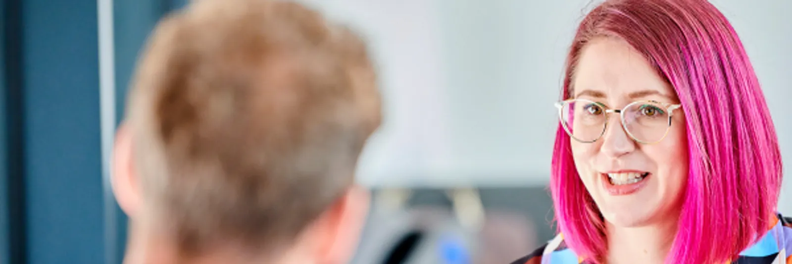 A man and woman sit opposite each other on low comfy armchairs. They are next to a large window which overlooks the wooden exterior of the Palatine Centre. He is wearing an open necked shirt and she is wearing a brightly patterned dress and has magenta pink coloured hair. They are talking and look as if they are having an informal meeting / catch-up