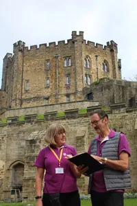 A woman and a man in purple uniform polo shirts stand in front of a historic stone wall and high stone building. The man holds a clip board and the two are talking.