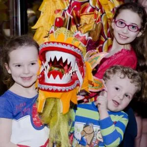 Four smiling children are holding up a brightly coloured Chinese parade dragon while standing next to a display case holding Chinese ceramics.