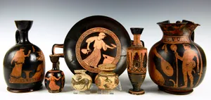 Seven pieces of Ancient Greek ceramics, on a graduated background. Ceramics include a wine cup, oil containers, and ointment pots.