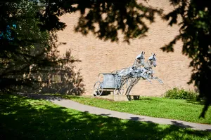 A life-size metal sculpture of a two-wheeled chariot pulled by two rearing horses stands on a lawn close to a path. Behind is a brick wall.