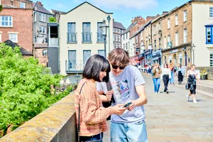 A pair of students looking at a phone screen together while stood on Elvet Bridge in Durham, with other people walking along in the background