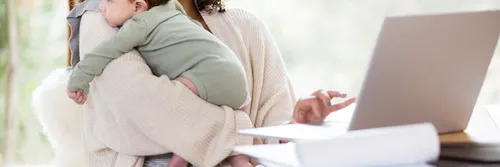 New mother scrolls on laptop while seated at her desk whilst holding her baby.