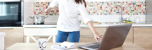 Woman in white shirt and blue jeans working on laptop and cooking at the same time
