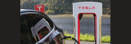 A close up of the rear wing of a black Tesla car as it charges at a red and white Tesla branded charging station.