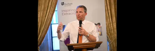 A man in a shirt and tie, sleeves rolled up, speaking into a handheld microphone
