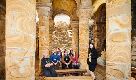 A group of people sit on wooden benches between stone columns in a room with stone walls. Guide stands to one side speaking.