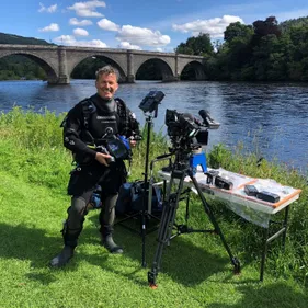 Image of Gary Bankhead, underwater archaeologist standing with filming equipment in front of a river.