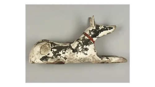 Ancient Egyptian Anubis statue depicting a seated dog painted black which has flaked away revealing the white clay underneath. The dog has long pointed ears and  a red collar around its neck.