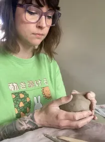 Artist with purple glasses and shoulder-length dark hair, in a green shirt, holding a lump of clay.