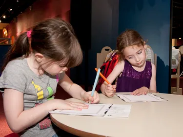 Two children sit at a table drawing a picture with coloured pencils.