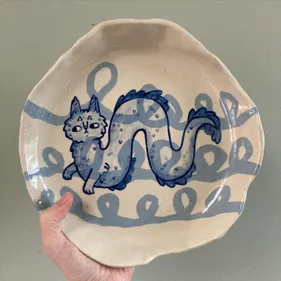 Hand holding hand made ceramic plate with blue hand illustrated cat like lambton worm