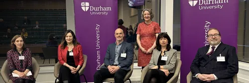 A mixed group of men and women sitting down. A women in a red dress stands behind them in between two purple 'Durham University' banners