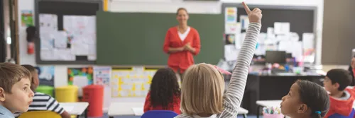 A young girl raises her hand to get a teacher's attention in a primary school classroom