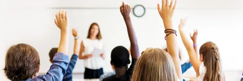 A group of school pupils with their hands up in a classroom with a teacher.