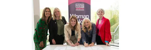 Five women standing behind a table. Two are leaning over the table, holding pens over a document