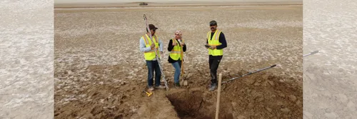 Two male and one female archaeologists stand by a deep trench in a desert landscape