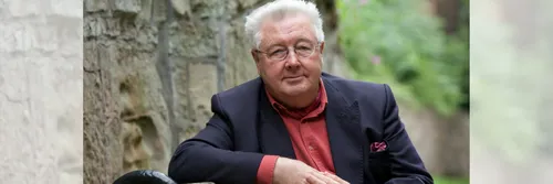 Douglas Davies sitting outside wearing a nave blazer and red shirt