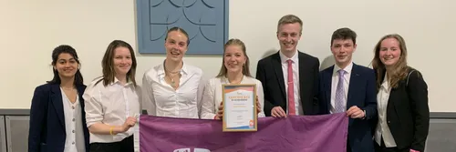 Group of smartly dressed students standing in a row smiling at the camera. The woman in the middle is holding a framed certificate.