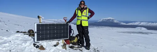 Dr Jenny Jenkins in a hi-vis vest stood in a snowy Icelandic landscape resting her hand on seismology equipment