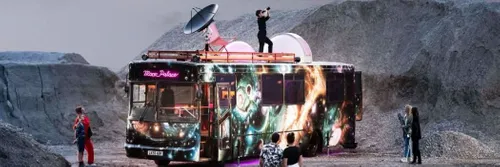 A single decker bus spray painted with images of space and featuring a satellite dish on its roof is parked in a dusty, moon-like landscape.
