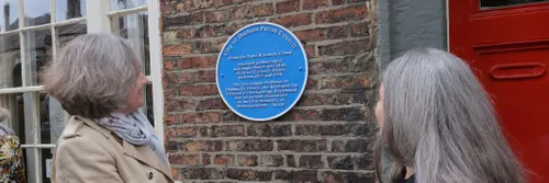 Professors Karen O'Brien and Sarah Semple look at a round blue plaque, set against a red brick wall, in honour of Professor Dame Rosemary Cramp.