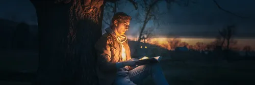 A man in a jacket sits under a tree in the dark reading a book, with a light illuminating his face