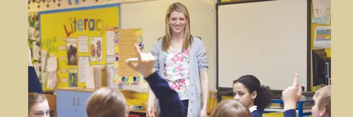Female teacher smiles at a class of children with their arms raised