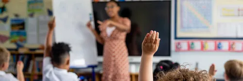 A child with their hand raised in a classroom with a teacher in the background in front of a blackboard