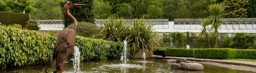 A heron sculpture sits in a pond at the Botanic Garden with greenhouses and plants in the background