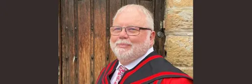 Professor Jon Gluyas with white hair, beard and glasses stands in front of an old wooden door in red and black academic robes.