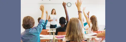 A teacher stands in front of a white board while students sitting in front of her raise their hands