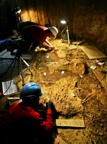 Three archaeologists in protective gear excavating an ancient burial site, with aid from large lamps.