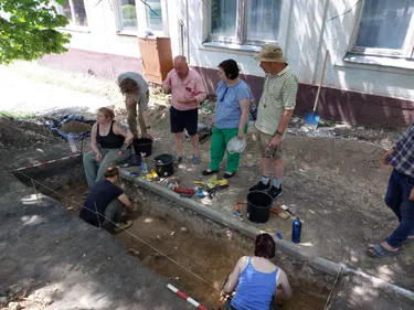 Three people stand next to an archaeological trench that is being excavated by Durham undergraduate students. Remains of stone tools are dotted around the trench centre and there is also evidence that mammoth tusk was worked.