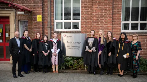 Group photo of 10 smiling people in formal dress standing outside a red brick building, spaced evenly around a metal building sign reading 'Durham University Anthropology Archaeology Dawson Building'. 8 of the 10 are also wearing black graduation gowns.