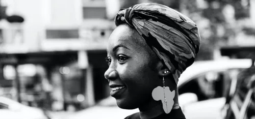 Monochrome photo of a Black woman standing side on to the camera, smiling and wearing an earring and headscarf. In the background is a busy city street.