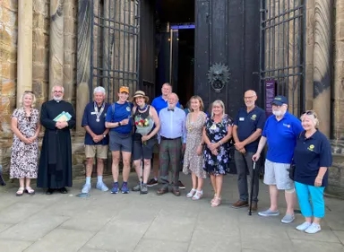 A group of smiling people gathered outside the Durham Cathedral doors. Two in the centre are dressed in hiking gear and one holds a bouquet of flowers.