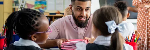 A male teacher smiling at two primary school pupils sitting at a desk in a classroom