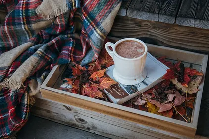 A mug of hot chocolate on a book in a tray full of autumn leaves, next to a blanket-covered chair
