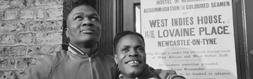 Two West Indian Merchant Seamen stand outside the 'West Indies House' hostel at 14-16 Lovaine Place, Newcastle-Upon-Tyne, 1941. Behind them can be seen a poster advertising the hostel, its location and facilities. The poster also states that the name of the superintendent is Mr Larbi.