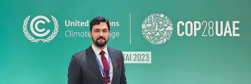 A man with a beard standing in front of a green sign displaying the COP28 logo.