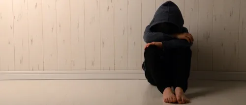 An upset child sitting against a wall with a hood pulled tightly over his head