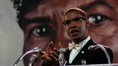 A still from the film, Malcolm X, showing a man speaking into a microphone against the backdrop of a face in the background