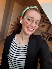 A portrait of a white female with curly red hair tied back, wearing a green and white headband, a pair of glasses, air-pods in her ears, a black and white stripy top and a black blazer
