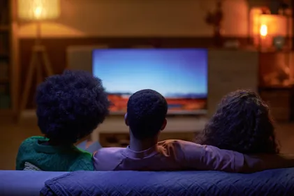 Photograph of three people sitting on a sofa watching the television, with atmospheric lighting and backs facing the camera.