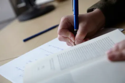 A student writing on a sheet of paper with a book open in the foreground