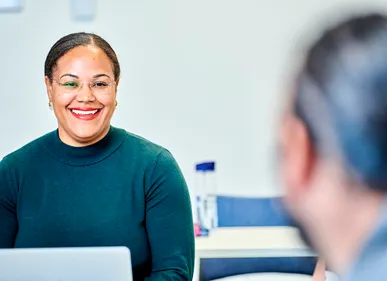 A woman with black hair pulled back off her face and clear framed glasses smiles at colleagues off camera. She wears a dark grey turtle neck jumper and is sitting inside a meeting room. She is an associate professor in the Sociology department